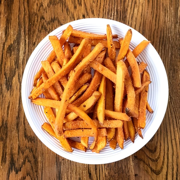 A white bowl filled with golden-brown sweet potato fries, seasoned with salt and pepper, on a wooden table.