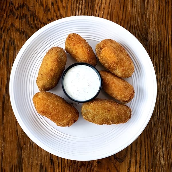 Six golden-brown fried appetizers, possibly kibbeh, arranged around a small cup of white dipping sauce on a white plate.